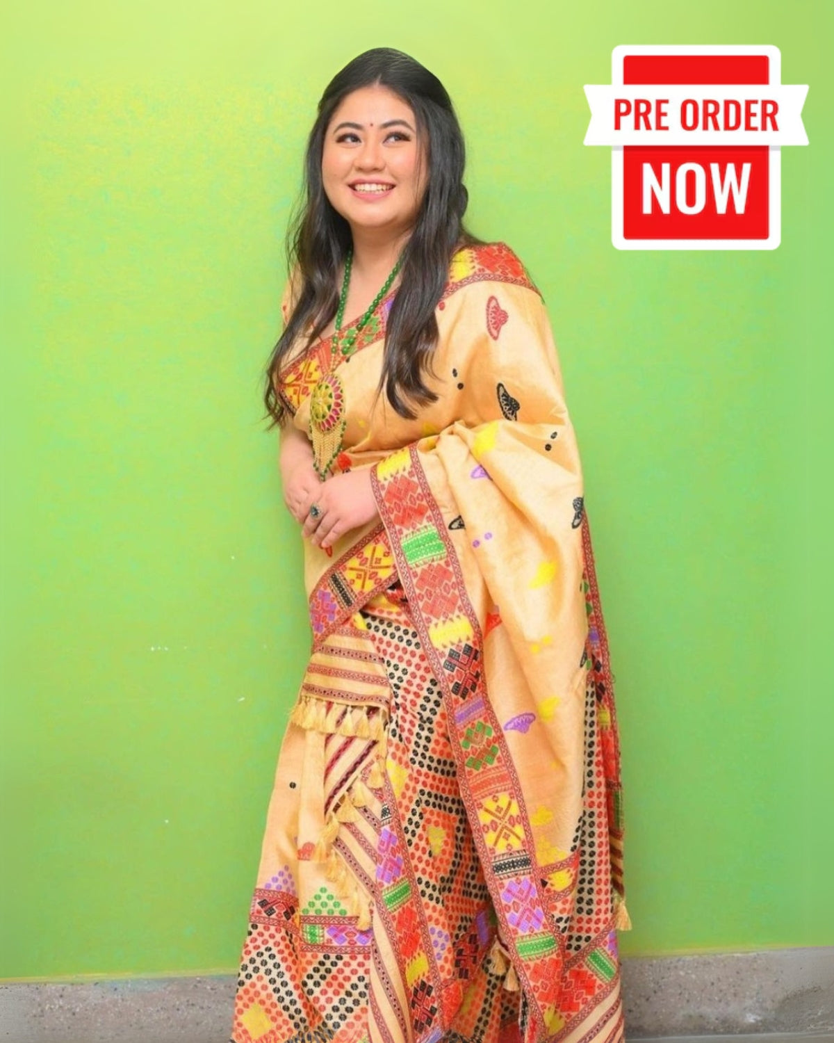 A woman standing against a green background, wearing a traditional Assamese mekhela sador with Mising tribal motifs in warm golden color.